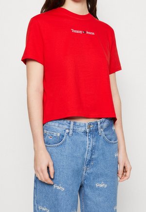 Kobieta ubrana w czerwony krótki T-shirt z logo "Tommy Jeans" oraz wysokiej talii niebieskie jeansy z haftowanymi naszywkami z napisem "Tommy Jeans".