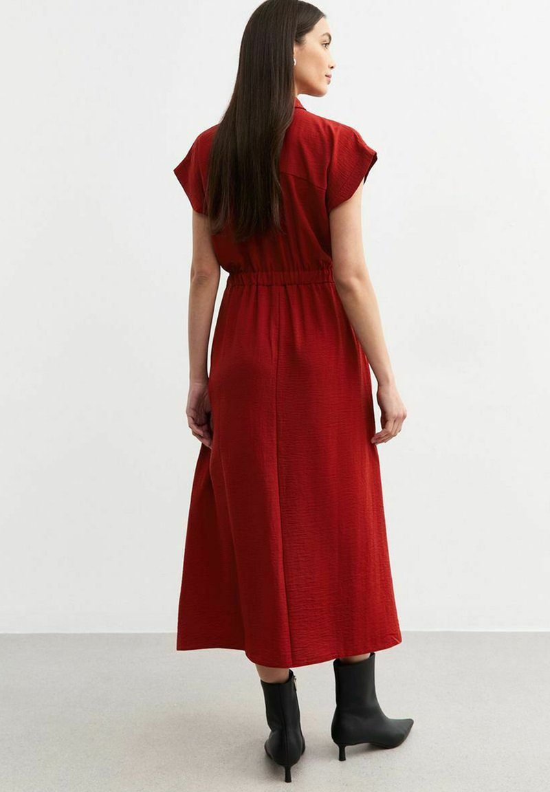 Vestido maxi rojo con mangas cortas, cintura ceñida, tejido texturizado y una silueta fluida, combinado con botas negras a los tobillos.