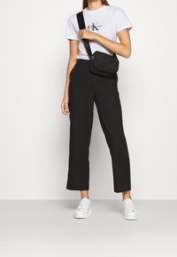 Černé kalhoty s vysokým pasem spárované s bílým tričkem s logem a bílými teniskami. Malá černá kabelka přes rameno dokresluje celý outfit.
