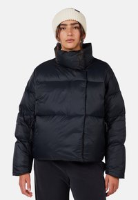 Giacca a vento nera con colletto alto e silhouette squadrata. Presenta polsini a coste e una finitura liscia e lucida. Abbinata a un berretto di maglia color crema.