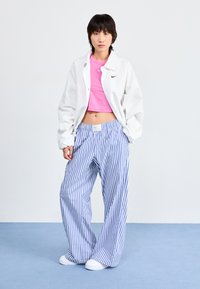 Bela oversized jakna, ružičasti crop top i široke hlače s plavim prugama. Manekenka nosi bijele tenisice, stoji na svijetloplavoj površini.