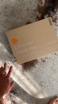 Brun kartonglåda med vit text "Älska mig. Bär mig." och en orange logotyp. Lådan har grova kanter och står på en texturerad yta.
