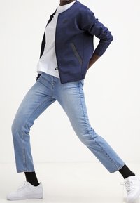 Marinblå zip-up jacka med grå accenter, vit t-shirt med rund hals, ljusblå jeans och vita sneakers med svarta strumpor. Avslappnad outfit.