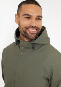 Veste étanche vert olive avec un col haut, des boutons-pression, un cordon de serrage réglable à la capuche et une texture lisse et brillante.