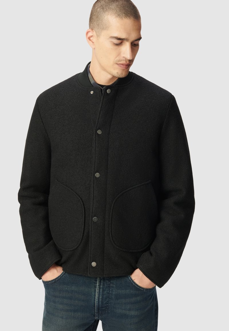 Czarna kurtka z mieszanki wełny z okrągłym kołnierzem, zapięciami na zatrzaski i dużymi przednimi kieszeniami. Teksturowany materiał o dopasowanym kroju i długich rękawach.