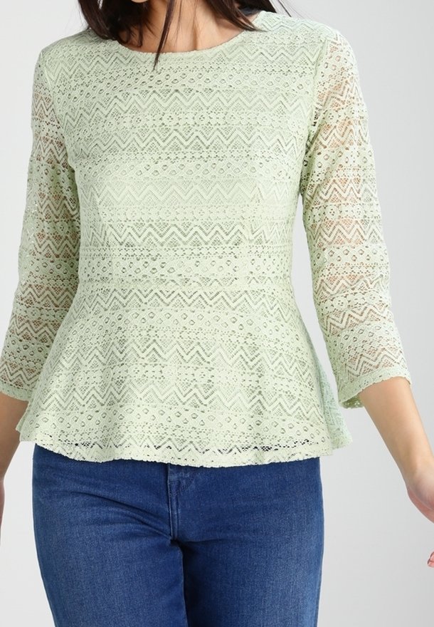 Femme portant un haut en dentelle vert pastel avec des manches trois-quarts et un bas à basque, associé à un jean bleu foncé.