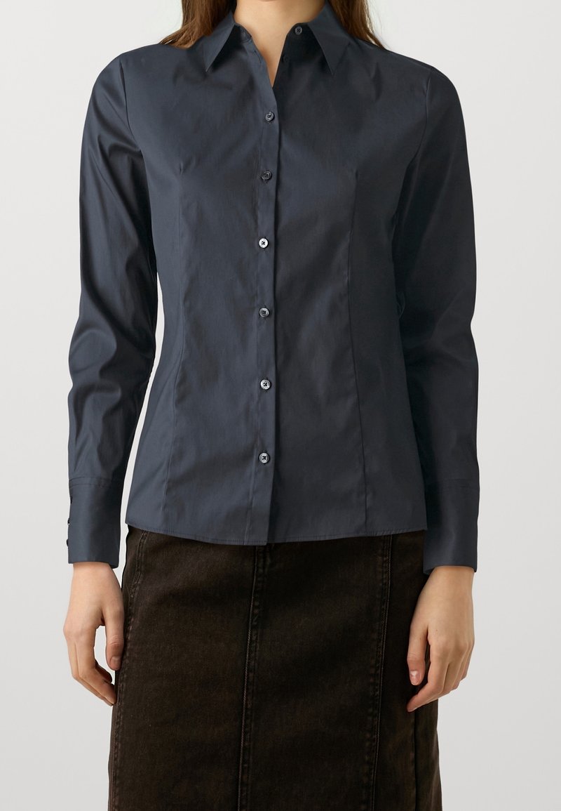 Femme portant une chemise bleu foncé cintrée à boutons avec des manches longues et une jupe marron foncé, debout devant un fond clair uni.