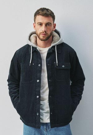 Man with short brown hair and beard wearing a black corduroy jacket with a grey hooded lining and blue jeans standing against a plain wall.