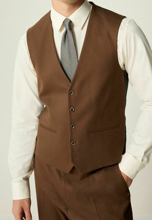 Homme portant un gilet marron boutonné et un pantalon assorti avec une chemise blanche et une cravate grise à motifs, une main dans la poche.