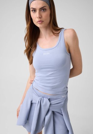 Femme aux longs cheveux bruns porte un débardeur bleu clair, une jupe assortie, un bandeau gris et un pull noué à la taille, debout à l'intérieur.