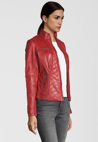 Rote Leder-Bikerjacke mit Stehkragen, gesteppten Schulter- und Seitendetails sowie silbernen Reißverschlussakzenten. Kombiniert mit einem weißen Hemd.