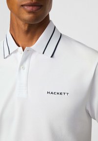 Biely polokošeľu s textúrovanou látkou, ktorá má kontrastný námornícky golier a manžety. Logo "HACKETT" v čiernej výšivke na hrudi.