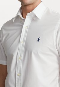Camisa blanca de botón con un collar clásico, con pequeños botones amarillos y un logo de jugador de polo en azul marino en el lado izquierdo del pecho. Textura de tela suave.
