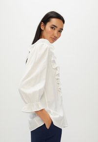 Blusa bianca con dettagli arricciati sul davanti, maniche a palloncino e polsini fronzuti. Realizzata in un tessuto leggero, presenta una vestibilità rilassata e un orlo dritto.