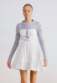 Vestido blanco de algodón con un corpiño ajustado, detalle de corte y cordón en la cintura; falda en capas; combinado con una blusa gris de mangas largas.