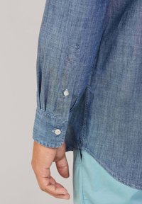 Lichtblauw denim shirt met een gestructureerde afwerking. Voorzien van knoopmanchetten en een gebogen zoom. Toont twee knopen op de manchetten.
