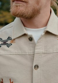 Beige shirt met een textuurstof, met intricaat borduurwerk in zwart, bruin en oranje patronen nabij de kraag. Twee knopen zichtbaar.