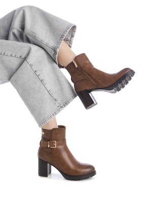 Botas de cuero marrón hasta el tobillo con un tacón grueso. Cuentan con una cremallera lateral, una hebilla decorativa y una suela texturizada para mayor tracción. Se combinan con pantalones de pierna ancha grises.