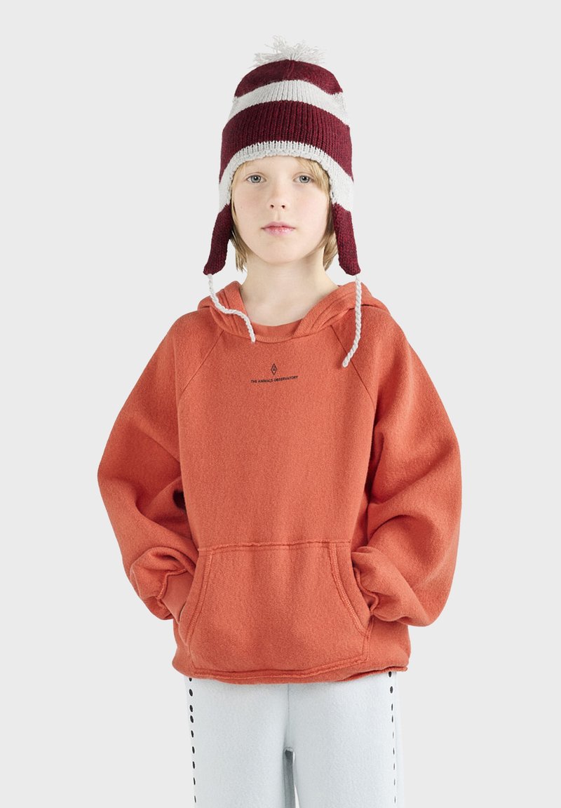 Dziecko w bordowej i białej w paski czapce z klapkami usznymi, pomarańczowym, oversize'owym hoodie oraz jasnych spodniach w czarne kropki.