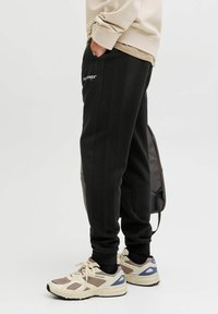 Zwarte joggerbroek met ribbelpanelen aan de zijkant, ritszakken, elastische tailleband en textuurstof, gecombineerd met beige sportschoenen.