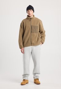 Giacca di pile marrone con zip frontale e tasca sul petto, abbinata a pantaloni sportivi grigi e stivali beige. Colori neutri con una vestibilità rilassata.