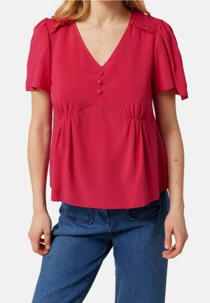 Kvinde iført en rød V-hals bluse med tre stofbetrukne knapper og puffede korte ærmer, kombineret med blå jeans.