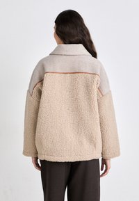 Giacca in pile beige con una texture morbida, spalle cadenti e accenti contrastanti color beige chiaro sul giogo e sulle maniche. Silhouette boxy.