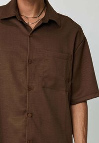 Chemise marron à manches courtes avec col, tissu texturé, poche avant et fermeture par boutons. Présente une coupe décontractée et un design simple.