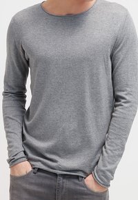 Maglietta grigia a maniche lunghe realizzata in tessuto morbido, con scollatura a girocollo e maniche aderenti, presentata con le mani in tasca su uno sfondo semplice.