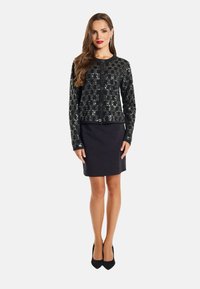 Cardigan nero con paillettes e motivo geometrico, con bottoni sul davanti, abbinato a una gonna nera attillata. Il modello indossa tacchi neri.