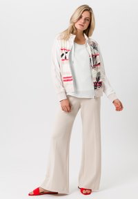 Veste blanche zippée avec un motif floral rouge et noir, associée à un débardeur léger et un pantalon ample beige, complétée par des sandales rouges.