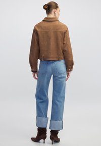 Veste cropped en daim marron avec col clouté, associée à un jean large bleu clair, avec des poignets effilochés et des bottes à talons marron.