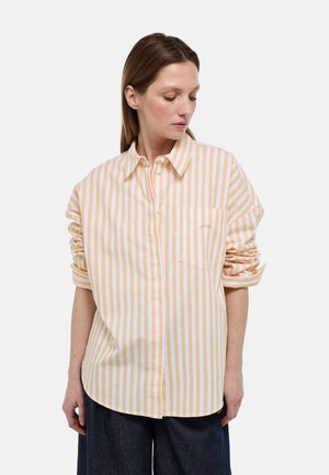 Femme portant une chemise ample à rayures verticales jaunes et blanches, manches retroussées, regardant vers le bas à sa droite.