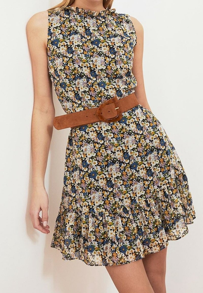 Robe sans manches à fleurs avec ourlet à volants, présentant de petites fleurs blanches, bleues et jaunes, cintrée à la taille par une large ceinture marron.