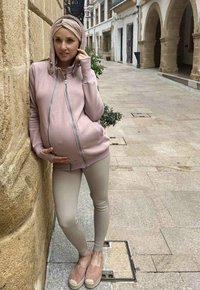 Felpa premaman rosa con zip e cappuccio, leggings beige e espadrillas rosa chiaro. Sfondo urbano con edificio in pietra e pavimentazione.