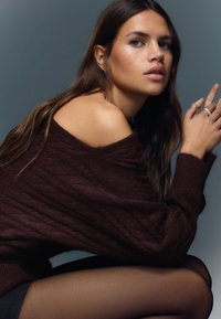 Dark brown oversized sweater with ribbed texture, worn off the shoulder. Black sheer tights complete the look. Soft-focus background.