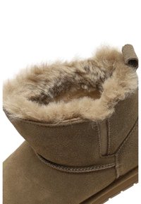 Bottine en daim marron avec une doublure en fausse fourrure beige douce. Présente un détail de couture et une languette en boucle à l'arrière.