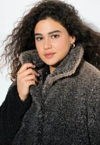 Rakenteellinen, suuri takki ruskean ja mustan sävyjen väreissä. Varustettu korkealla kauluksella ja napituskiinnityksellä. Pehmeä, pörröinen materiaali.