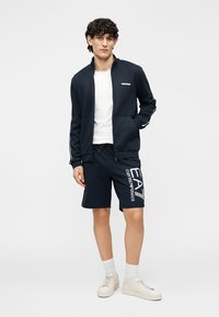 Tummansininen vetoketjullinen takki, jossa on kapeahko istuvuus, yhdistettynä yhteensopiviin tummansinisiin shortseihin, joissa valkoinen "EA7 Emporio Armani" -painatus. Tennarit ja vaaleat sukat.