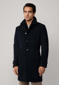 Uomo che indossa un lungo cappotto nero con bottoni, colletto ampio e tasche laterali, in piedi contro uno sfondo chiaro e semplice.