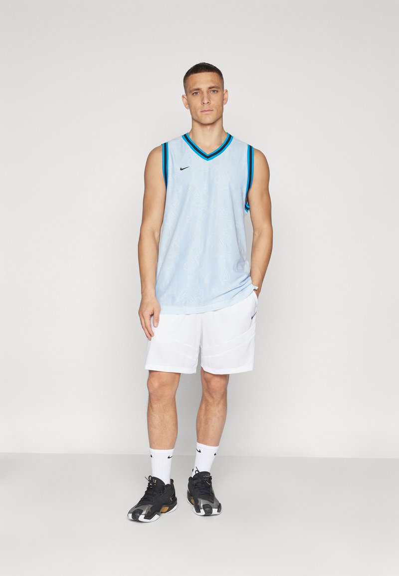 Maillot de basket sans manches bleu clair avec texture à motifs, logo Nike noir et bordure teal contrastante, associé à un short blanc et des chaussures noires.