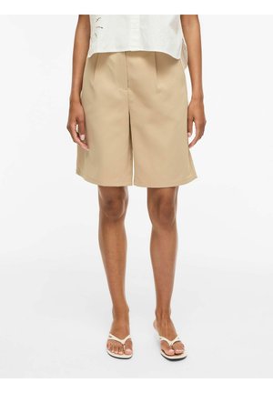 Person som har på seg beige shorts til knærne, hvit topp og hvite sandaler, står på en ensfarget hvit bakgrunn.