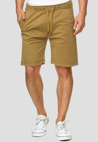 Shorts en coton beige avec une taille élastique, un cordon de serrage et des poches latérales. Texture lisse et coupe droite, se terminant juste au-dessus du genou.