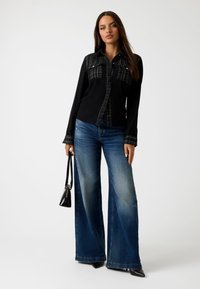 Zwarte button-up blouse met gestructureerde schouders, gecombineerd met lichtblauwe brede jeans. Model houdt een kleine zwarte handtas vast.