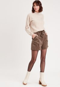 Pull en tricot beige avec motif en câble, associé à un short en velours côtelé marron. Le modèle porte des bottes à talons blanches et des collants noirs transparents.