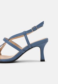 Sandale en cuir bleu avec des brides croisées, boucle de cheville réglable et un talon carré de 6,5 cm. Texture lisse et design minimaliste.