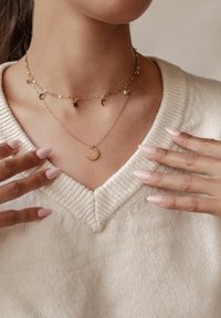 Collarín de oro con charms de estrella y luna; cadenas finas; suéter texturizado de color crema claro; uñas manicuras de color rosa pálido.