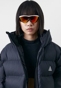 Black puffer jacket with a high collar and quilting details, layered over a ribbed black sweater. Large reflective sunglasses with orange lenses.