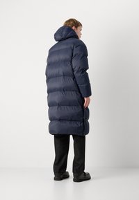 Marineblå puffjakke med hette, med en lang, quiltet design og en glatt, skinnende tekstur. Inkluderer en sidesplitt, brukt med mørke bukser og sko.