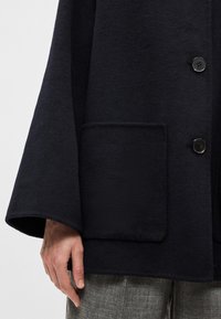 Tmavě námořnický vlněný kabát s volným střihem, širokými rukávy a přední kapsou. Má zapínání na jeden knoflík a měkkou texturu.